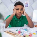 Meditation. Pensive dark-skinned boy of primary school age in green tshirt touching head with hands looking aside sitting at table with drawing and brushes in daylight